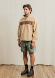 Man wearing a beige sweater with a brown stripe, green shorts, and black boots standing on a wooden floor.