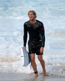John John Florenece in a F1 Standard Issue Boardshort holding a surfboard in shallow water with waves in the background