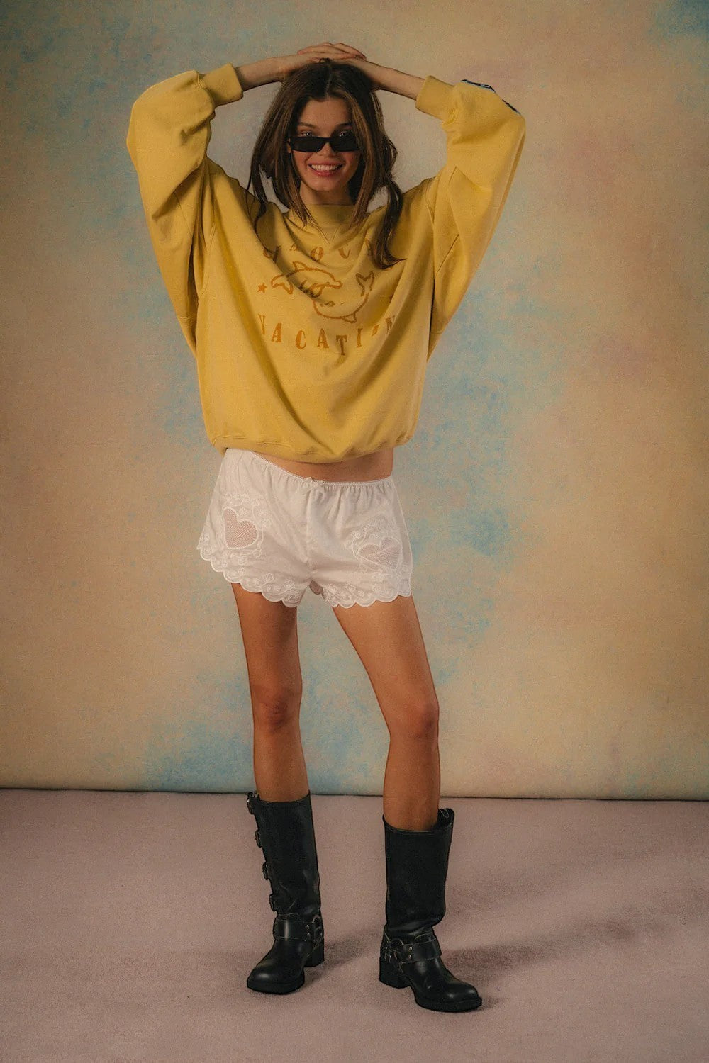 Keel Surf & Supply yellow Delfino sweater with dolphin graphic and "Vacation" text, worn by a model with white lace shorts and black boots.