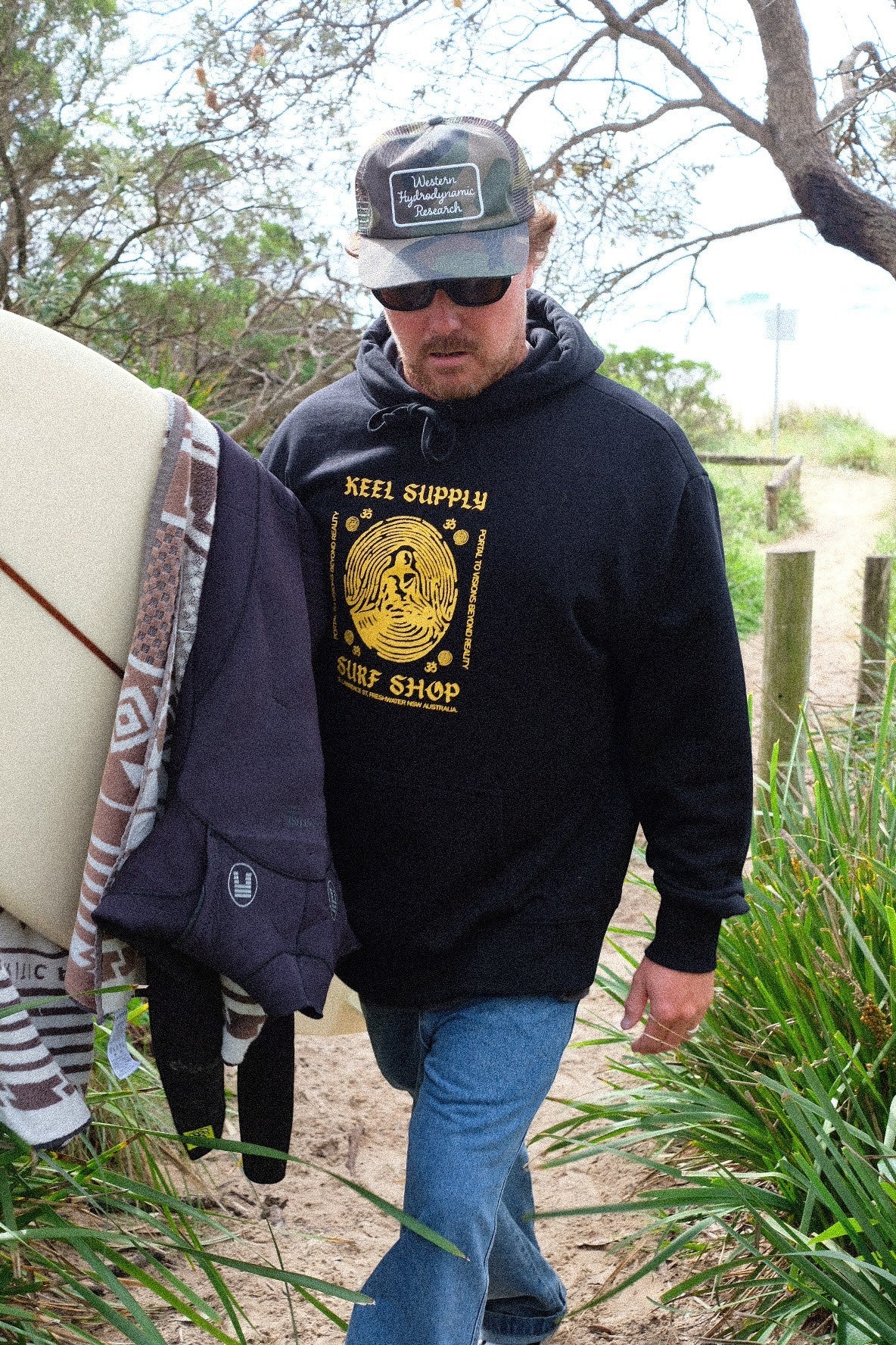 Keel Supply black heavyweight fleece hoodie with yellow meditation graphic and pullover hood. Worn by a man carrying a surfboard.