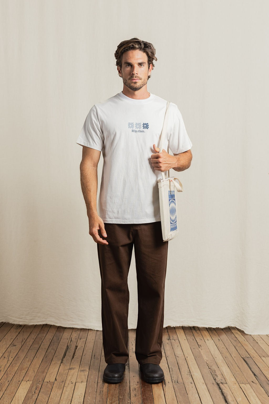 Keel Surf & Supply white cotton t-shirt with blue graphic print and "Rhythm." text. Model wears brown pants and carries a tote bag.